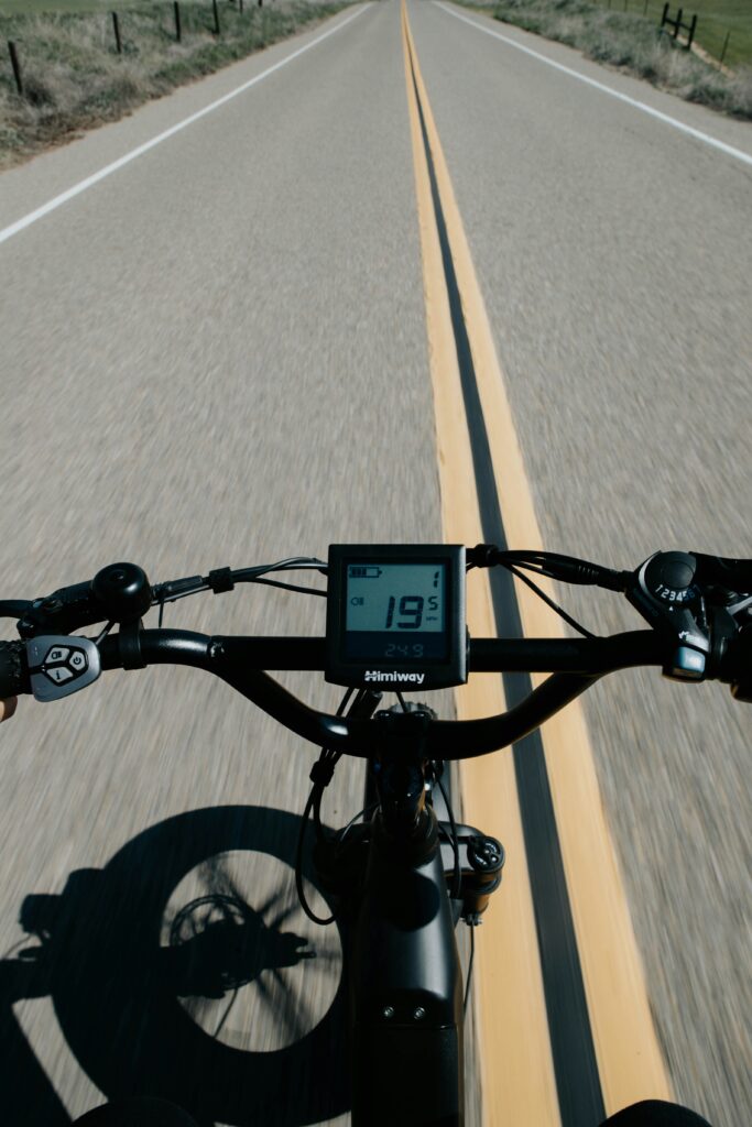 The handlbars of an e-bike riding down a road, showing its speedometer at 19 miles per hour